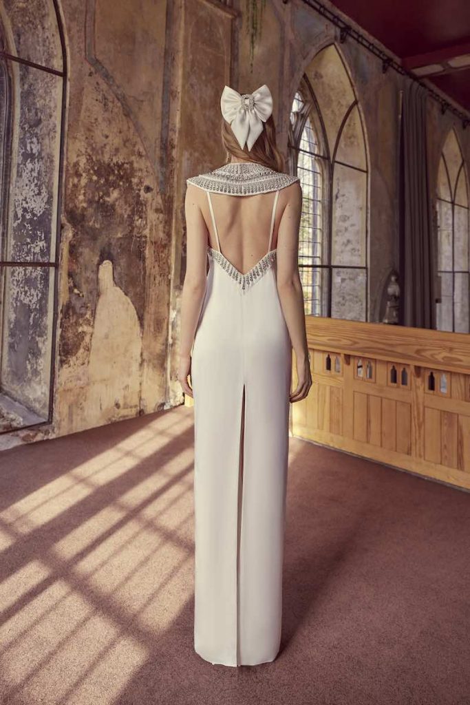 In questa foto una modella di spalle  indossa un abito da sposa Temperley Bridal 2024 in crêpe con vollatura sulla schiena decorata sul bordo con paillettes. La modella indossa anche un fermaglio a forma di fiocco abbinato