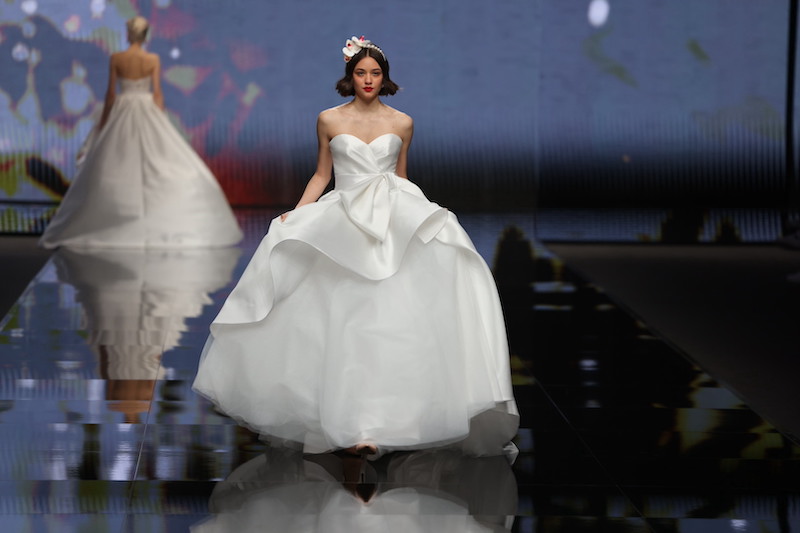 In questa foto una modella sfila indossando un abito da sposa Mysecret 2024 con gonna in organza e sopragonna in mikado con maxi fiocco frontale