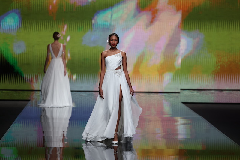 In questa foto una modella sfila indossando un abito da sposa scivolato con gonna i chiffon con spacco, corpetto monospalla e dettaglio cut-out sul fianco sinistro