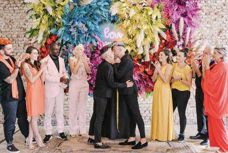 In questa foto due sposi con i colori invitati si baciano davanti ad un backdrop con fiori dai colori fluo azzurro, giallo, fucsia e rosso