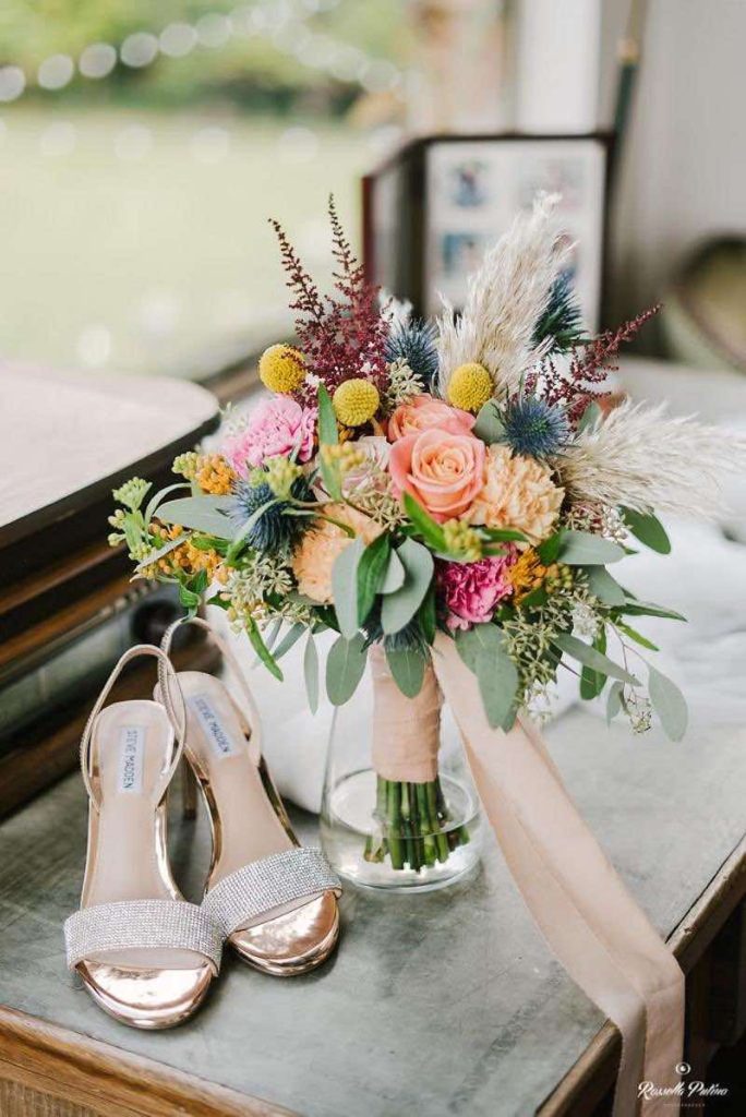 In questa foto il dettaglio di un bouquet da sposa realizzato da Idee da Sogno con fiori di colore rosa, rosa pesca, giallo e azzurro e  fotografato accanto ad un paio di sandali di colore oro e argento di Steve Madden