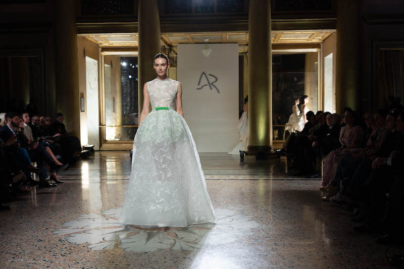 In questa foto una modella sfila indossando un abito da sposa Antonio Riva 2024 con gonna dalla linea ad A in tulle ricamato con corpetto girocollo e maniche corte. In vita, l'abito presenta un cinturino verde