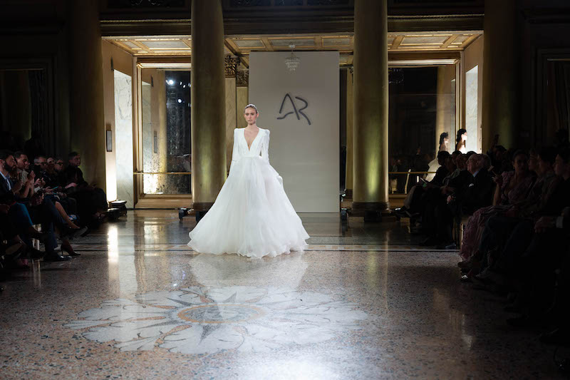 In questa foto una modella sfila indossando un abito da sposa Antonio Riva 2024 ampio in tulle, scollo a V profondo e maniche lunghe