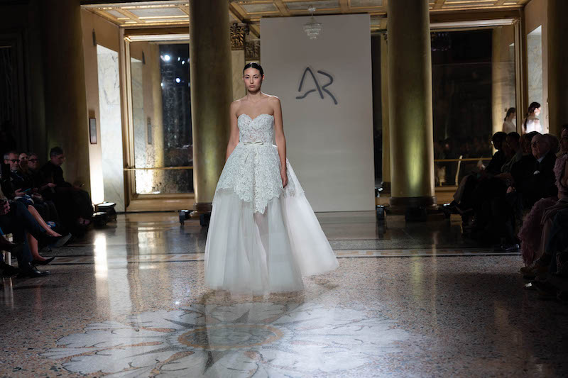 In questa foto una modella sfila indossando un abito da sposa Antonio Riva 2024 dalla linea ad A con corpetto con scollo a cuore in pizzo che scende fino a metà della gonna in organza e micro fiocco in vita