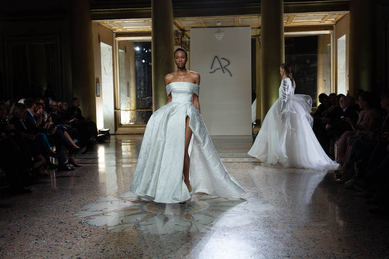 In questa foto una modella sfila indossando un abito da sposa Antonio Riva 2024 in broccato di seta con ampio spacco sul lato sinistro e scollo dritto off-shoulder