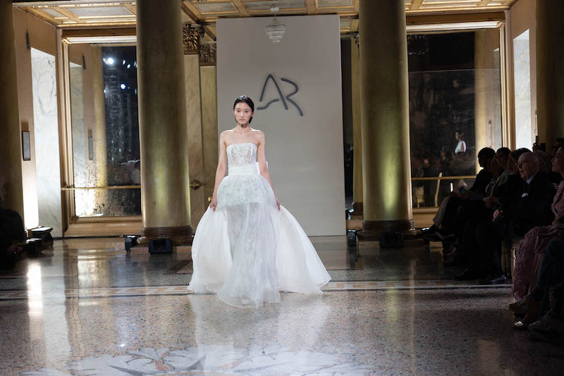 In questa foto una modella sfila indossando un abito da sposa Antonio Riva 2024 scivolato in pizzo con sopragonna abbinato, corpetto con scollo dritto e ampia cintura in vita che si chiude con maxi fiocco sul retro