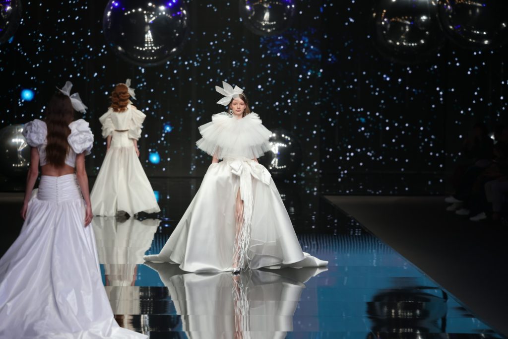 In questa foto una modella sfila indossando un abito da sposa Elisabetta Polignano 2024 della collezione Timeless con gonna in mikado di seta, spacco frontale fermato da un maxi fiocco in rafia. Il corpetto è coperto da una maxi cappa di tulle plissettato a balze. La modella indossa anche un cerchietto tra i capelli con maxi fiocco destrutturato