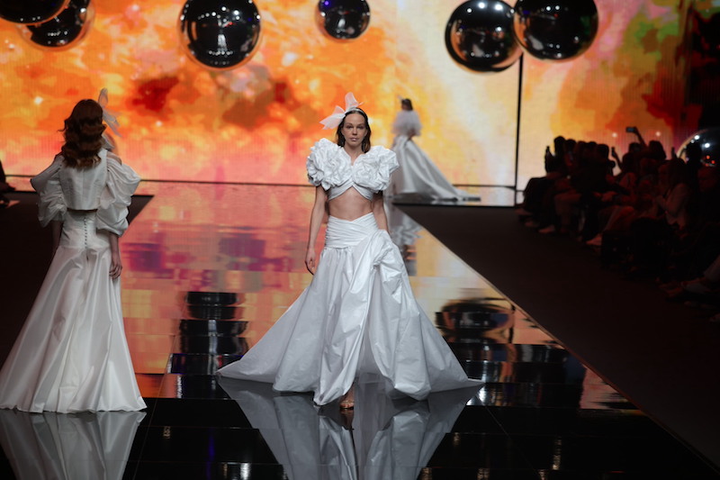 In questa foto una modella sfila indossando un abito da sposa Elisabetta Polignano 2024 composta da maxi gonna in taffetá e crop top abbinato con due maxi fiori che esplodono sulle spalle.La modella indossa anche un cerchietto tra i capelli con maxi fiocco destrutturato