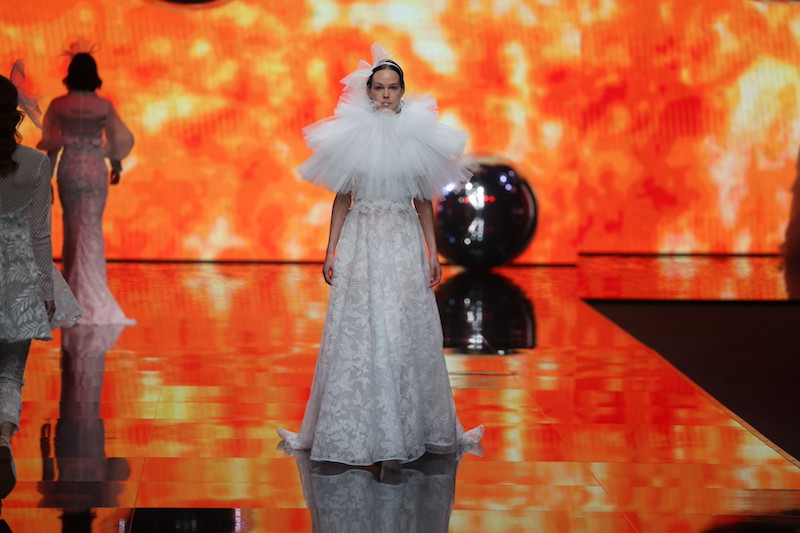 In questa foto una modella sfila indossando un abito da sposa Elisabetta Polignano 2024 in pizzo floreale dalla linea ad A con cappa di tulle plissettato che copre il corpetto. La modella indossa anche un cerchietto tra i capelli con maxi fiocco destrutturato