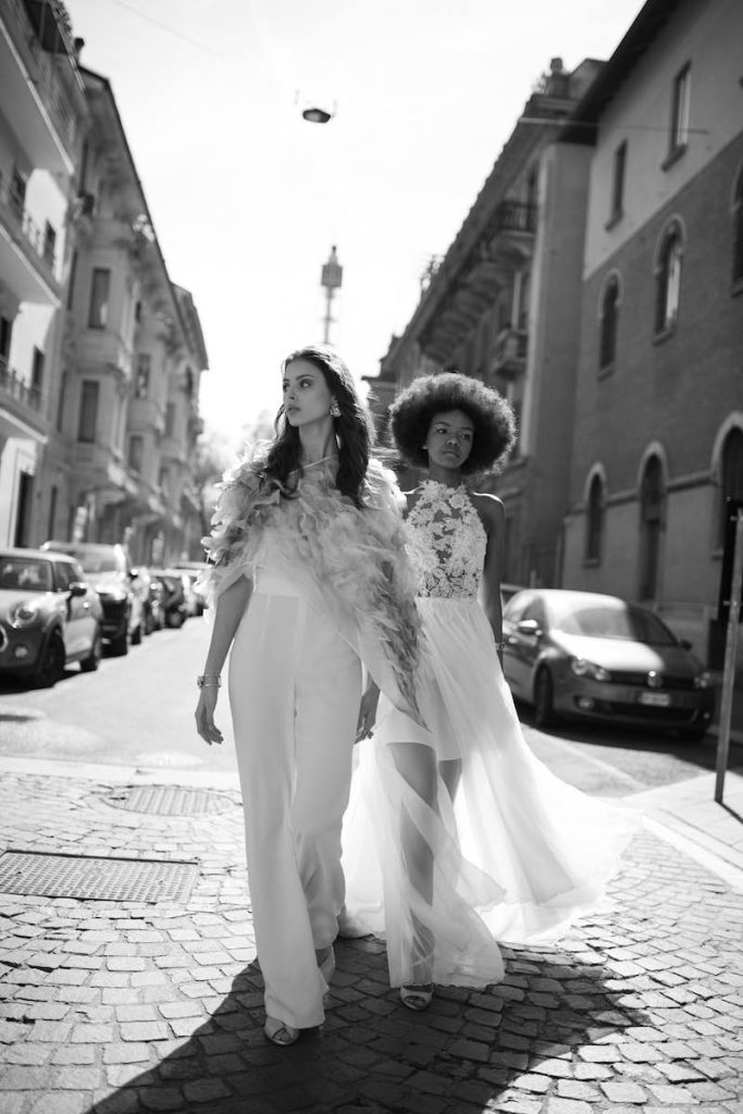 In questa foto in bianco e nero due modelle camminano per strada indossando una tuta da sposa con cappa plumage e un abito da spsoa scivolato con gomma in chiffon e corpetto in pizzo floreale de Le Fate Milano