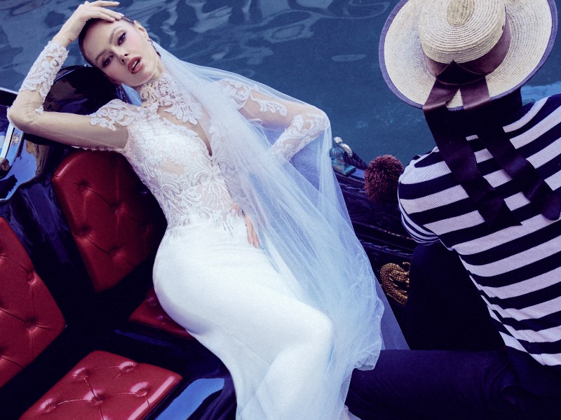 In questa foto una modella sdraiati all'interno di una gondola indossa uno degli abiti da sposa Maison Signore 2024 a sirena in pizzo, maniche illusion e velo abbinato