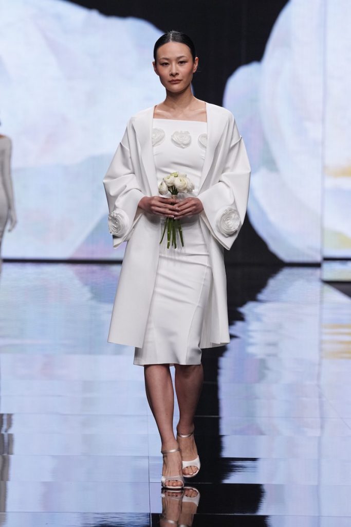 In questa foto una modella sfila indossando un abito da sposa a tubino di Chiara Boni abbinato ad un cappotto da sposa