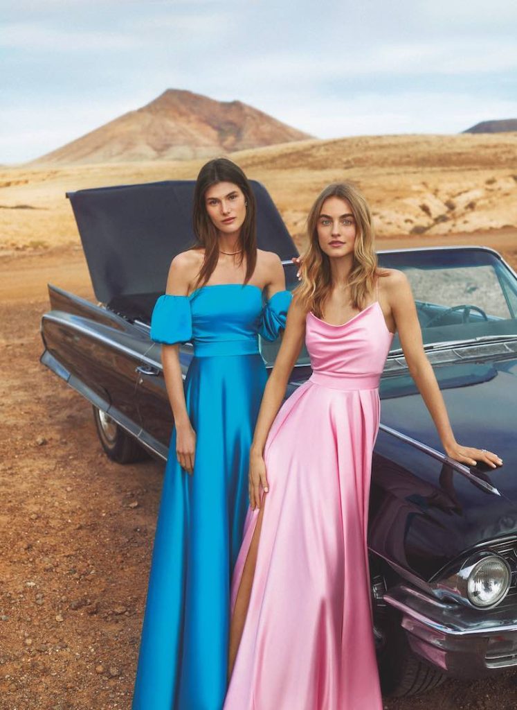 In questa foto due modelle appoggiate ad un'auto decappottabile vintage indossano due abiti lunghi da damigella Atelier Emé 2023 di raso satin nei colori turchese e rosa baby