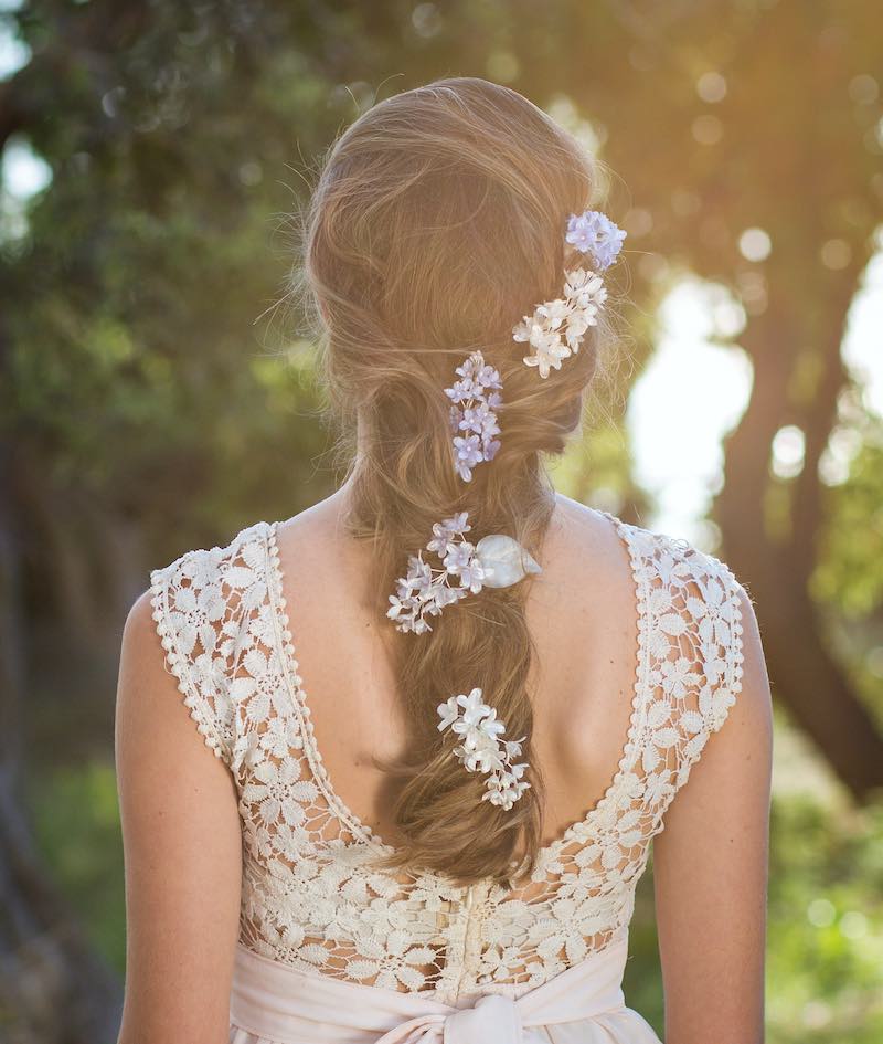 In questa foto una modella di spalle con i capelli acconciati in una treccia decorata con piccoli fiori di seta di colore lilla e bianco
