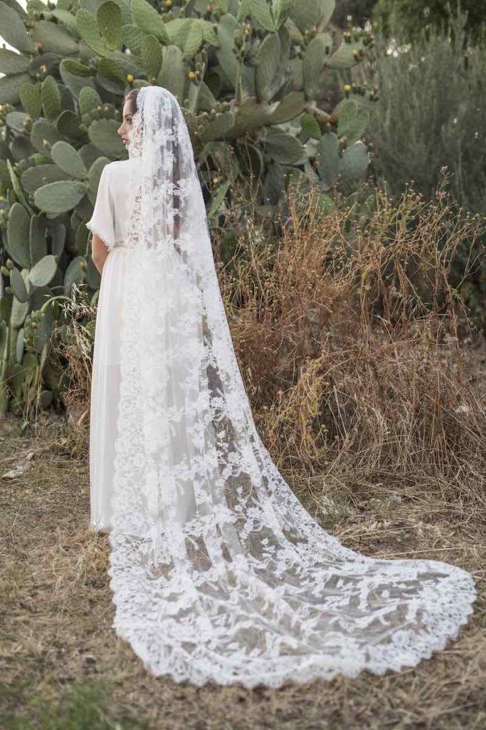 In questa foto una modella di spalle in un prato indossa un abito da sposa semplice e un velo di pizzo di Marco Cucinotta