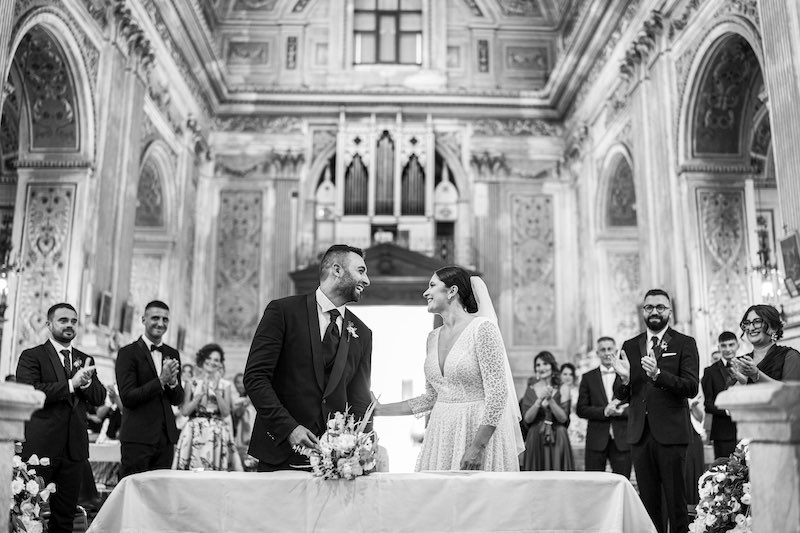 In questa foto in bianco e nero due sposi si guardando sorridendo dietro al banco posto all'altare. Dietro di loro amici e parenti battono le mani ed è mostrata tutta la chiesa per il matrimonio