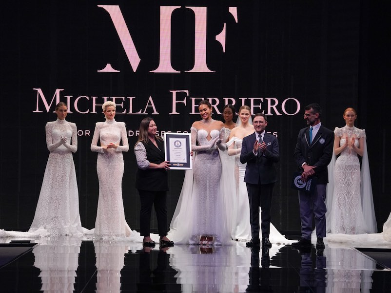 In questa foto la stilista Michela Ferriero riceve l'attestato del Guinness World Record sulla passerella del Sì Sposaitalia con il marito e ceo dell'azienda Lorenzo Albanese circondati da modelle che indossano gli abiti da sposa 2024 della collezione Enchanté