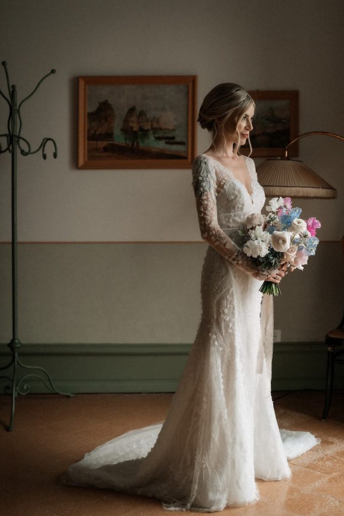 In questa foto Nina Rima posa davanti ad una finestra indossando il suo abito da sposa a sirena in tulle con: ricami 3D ad onde, maniche, scollo a V e strascico. Tra le mani tiene il suo bouquet da sposa con fiori bianchi, rosa e azzurri
