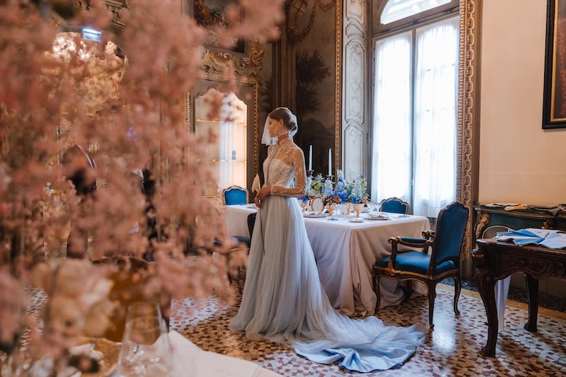 In questa foto una modella posa in abito da sposa di colore azzurro polvere in un salone nobiliare di Milano allestito con gypsophila di colore rosa e con un tavolo rettangolare decorato con fiori di colori blu