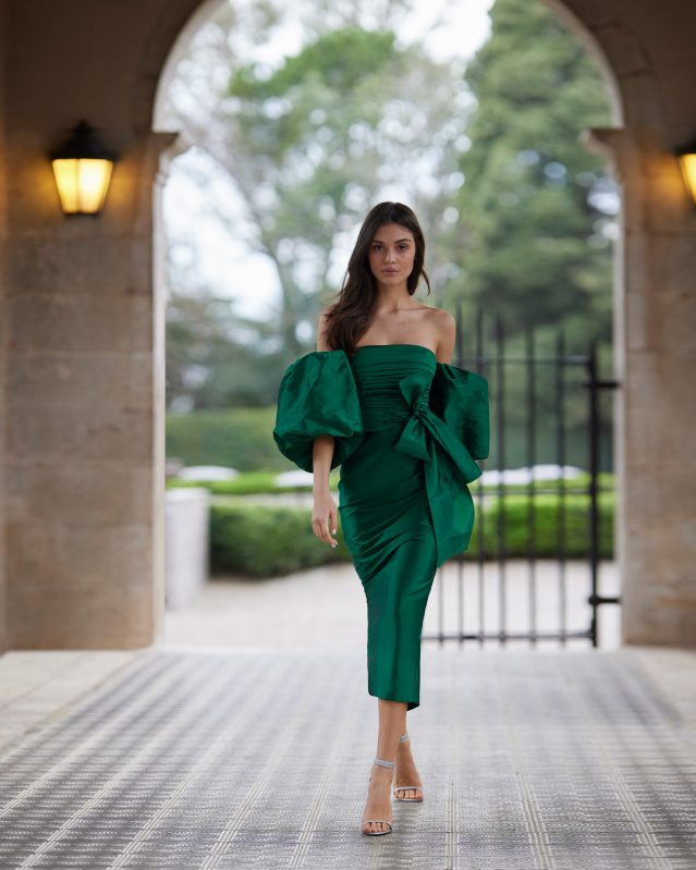 In questa foto una modella indossa un abito da cerimonia donna 2023 dritto midi, in stile classico, confezionato interamente in taffetà, con gonna stretta con spacco posteriore. L'abito è della collezione Rosa Clará cerimonia 2023, con scollo senza spalline, fiocco sulla schiena e maniche a sbuffo rimovibili