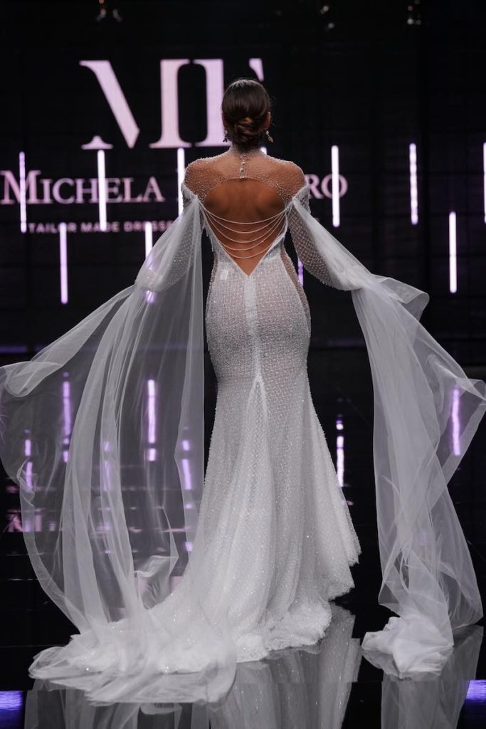 In questa foto una modella ritratta di spalle sfila al Sì Sposaitalia Collezioni 2023 indossando un abito da sposa a sirena di Michela Ferriero con ricami di cristalli Swarovski e maxi ali di tulle di seta