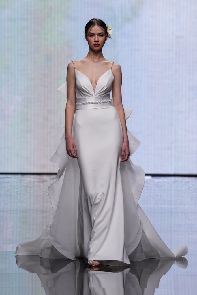 In questa foto una modella sfila al Sì Sposaitalia 2023 indossando un abito da sposa a sirena liscio in georgette con scollo a V, piccola cintura gioiello in vita e coda scultorea di Mysecret Sposa