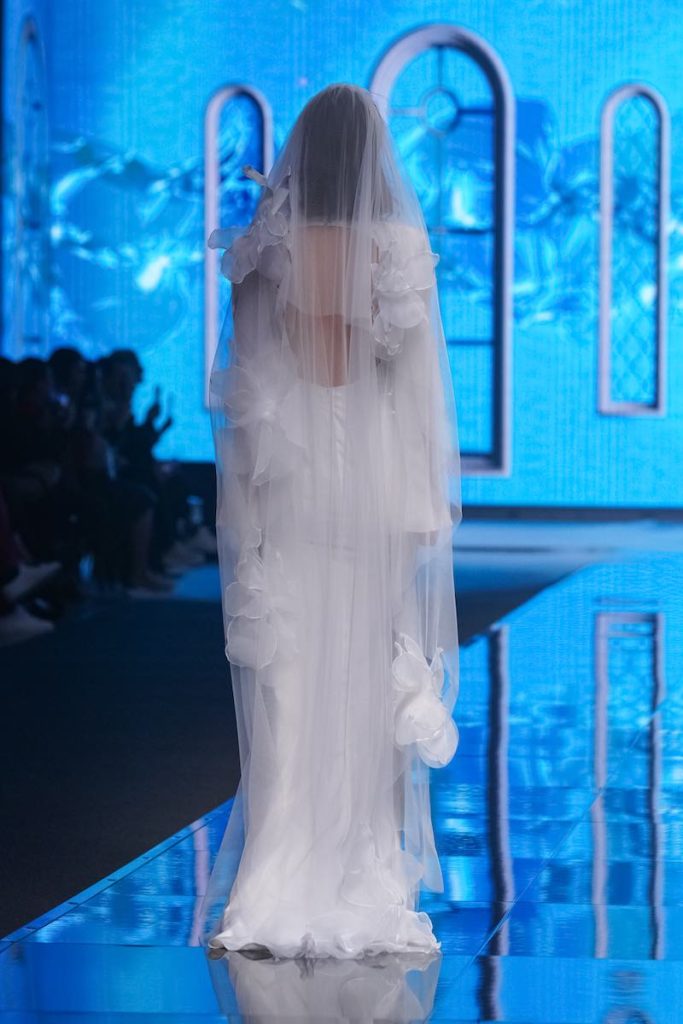 In questa foto una modella di spalle sfila indossando un abito da sposa con velo con maxi fiori 3D di Rara Avis