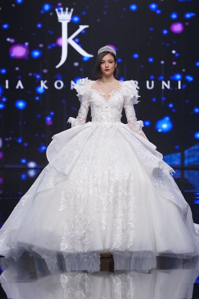 In questa foto una modella sfila al Sì Sposaitalia Collezioni 2023 indossando un abito da sposa principessa con ricami in pizzo, balze sui fianchi e pizzo in 3D sulle spalle di Julia Kontogruni