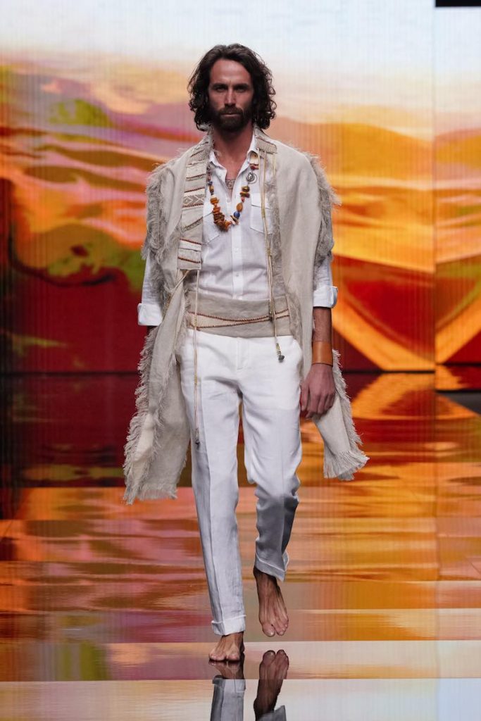 In questa foto un modello sfila indossando un abito da sposo stile boho chic composto da pantaloni e camicia di colore bianco, cintura in lino beige abbinata ad una stola di lino