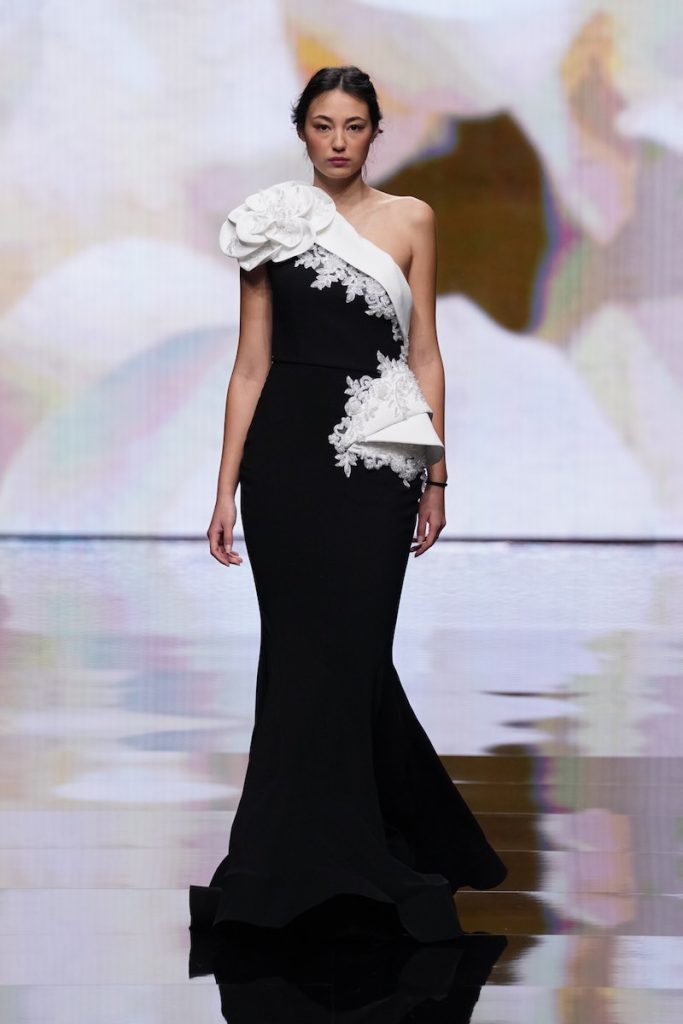 In questa foto una modella sfila indossando un abito da cerimonia di colore e nero e bianco, modello a sirena monospalla con ricami in pizzo e maxi fiore di colore bianco sulla spalla destra