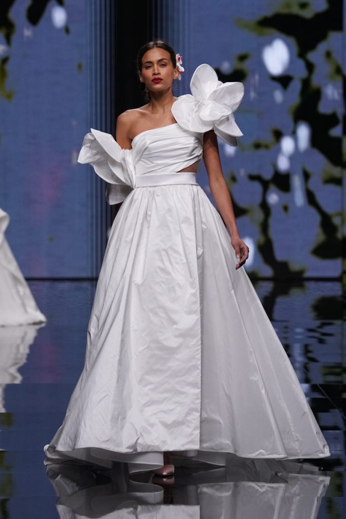 In questa foto una modella sfila al Sì Sposaitalia Collezioni 2023 indossando un abito da sposa monospalla in taffetà di Mysecret Sposa con gonna ad A, dettaglio cut-out sul fianco sinistro e maxi orchidee 3D sulle spalle