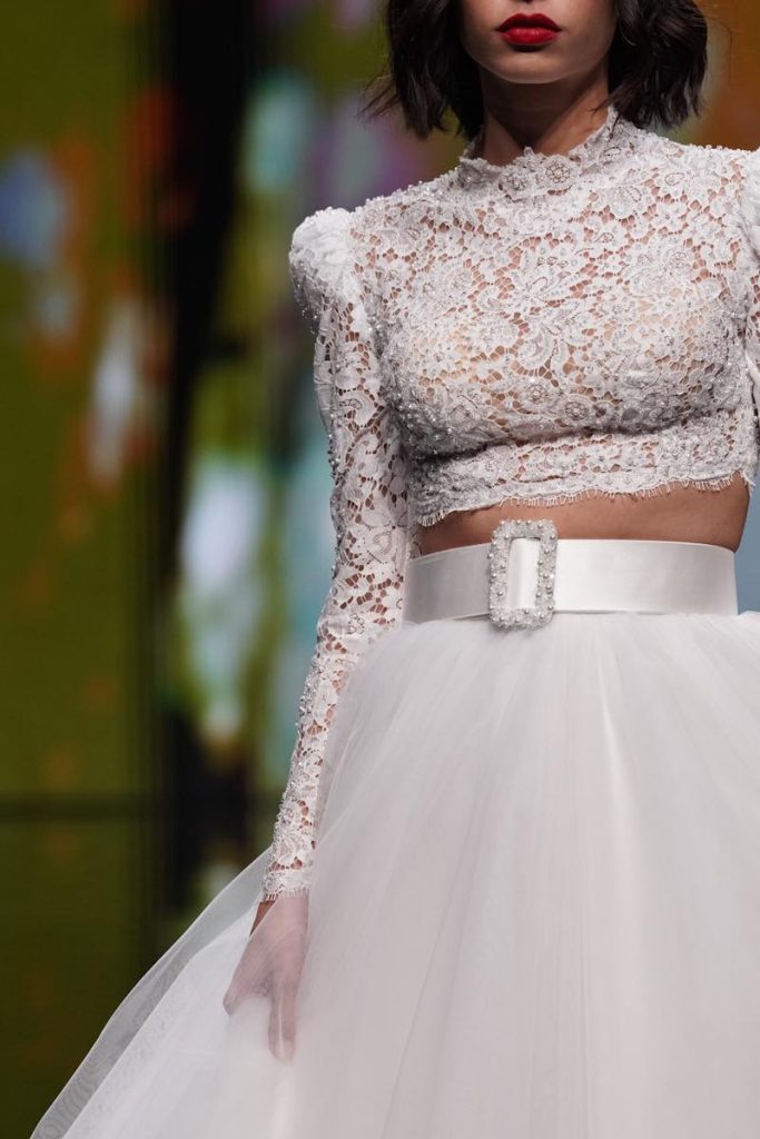 In questa foto una modella sfila indossando un abito da sposa due pezzi con gonna in tulle ampia fermata da un cinturone con fibbia gioiello abbinato ad un crop top in pizzo con maniche lunghe