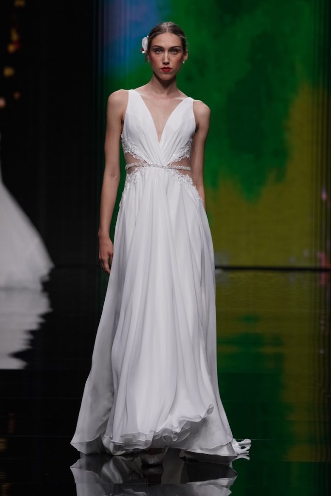 In questa foto una modella sfila al Sì Sposaitalia Collezioni 2023 indossando un abito da sposa in chiffon modello scivolato con dettagli cut-out sui fianchi con ricami di paillettes e periline