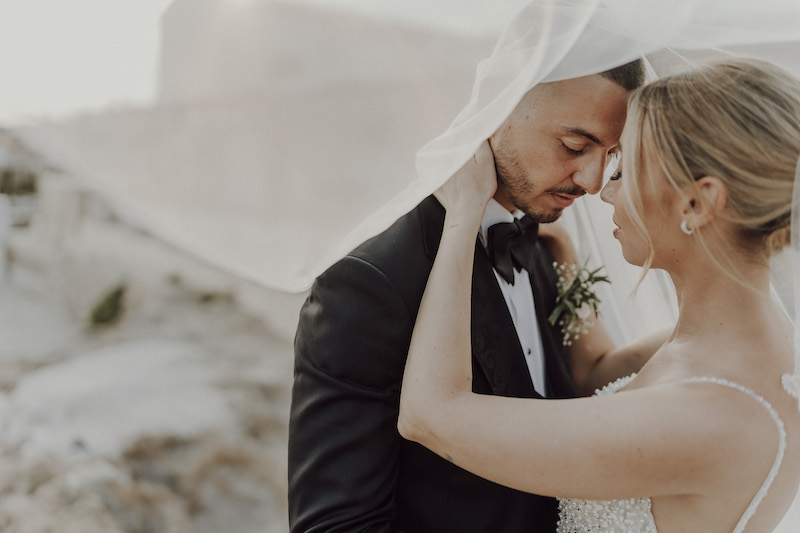 In questa foto due sposi si abbracciano sotto al velo di lei appoggiando la fronte l'uno sull'altra. La sposa tiene il viso dello sposo con la mano sinistra.