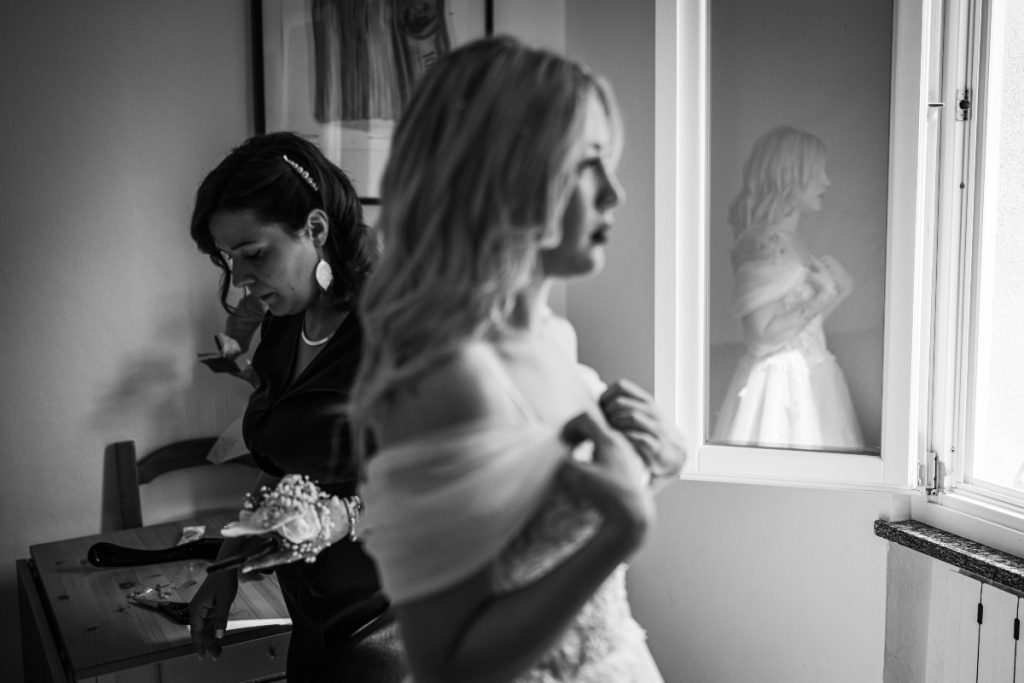 Una foto in bianco e nero di Riccardo Alù, tra i fotografi di matrimonio a Torino iscritti ad ANFM, in cui la sposa Johana si prepara ed il suo riflesso appare sulla finestra