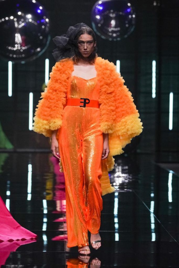 In questa foto un modella sfila indossando una tutta della collezione cerimonia Elisabetta Polignano 2024 di colore arancione fluo abbinata ad una mantella con ruches e un cinturone con monogramma 