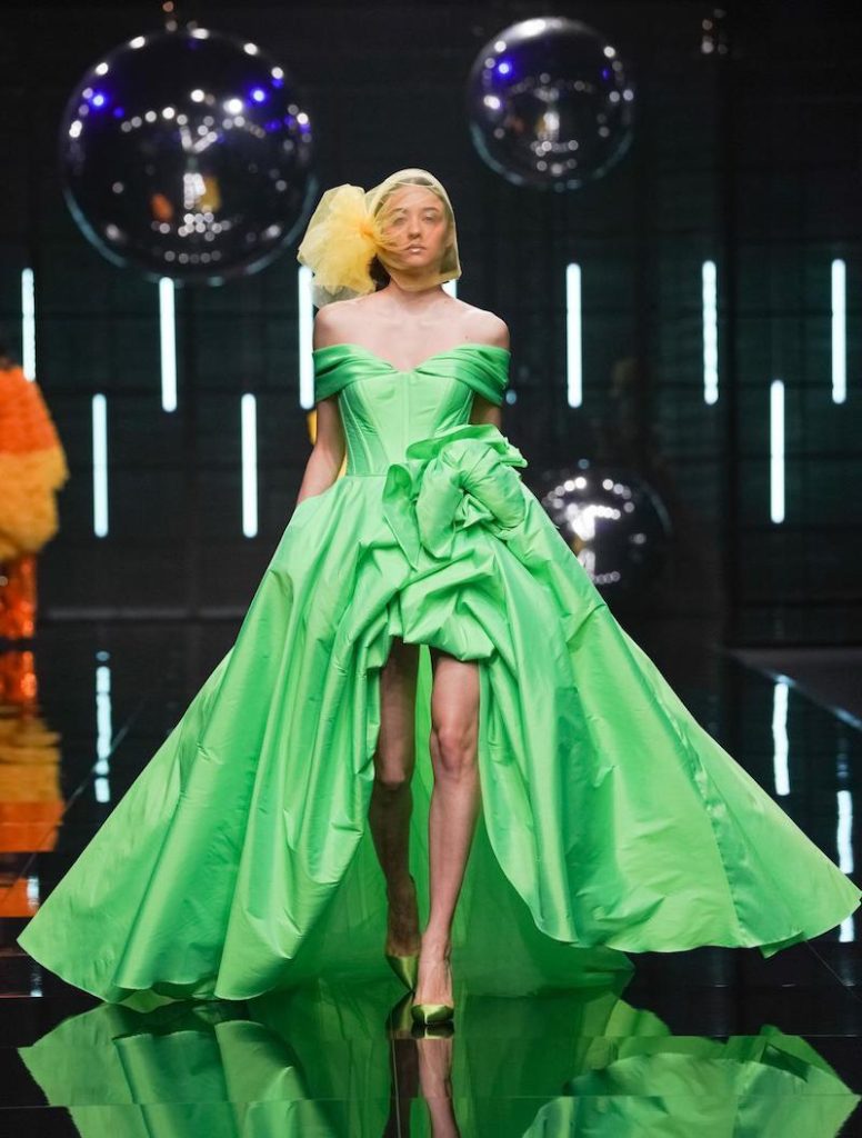 In questa foto una modella sfila indossando un abito da cerimonia corto davanti e lungo dietro di colore verde pistacchio e con un maxi fiore 3D in vita