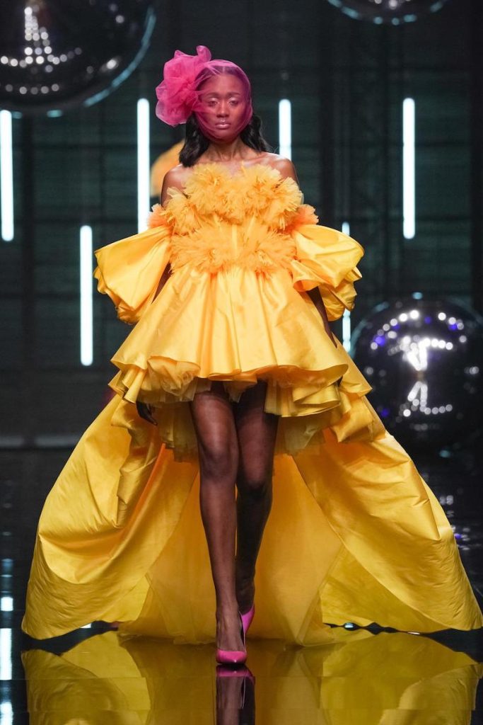 In questa foto una modella sfila indossando un abito da cerimonia Elisabetta Polignano 2024, in taffetà corto davanti e lungo dietro di colore giallo con maxi ruches sul corpetto