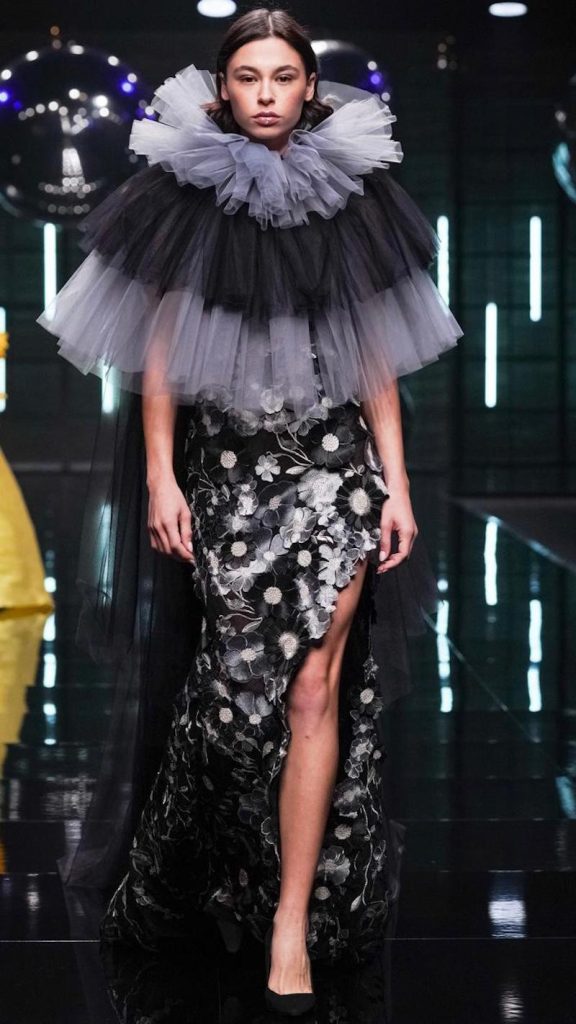 In questa foto una modella sfila indossando un abito da cerimonia modello a sirena con spacco laterale di colore nero con fiori 3D all over e un maxi collare di tulle di colore nero e grigio