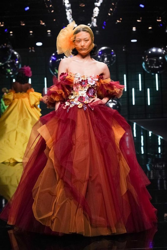 In questa foto una modella sfila indossando un abito da cerimonia di Elisabetta Polignano ampio di colore rosso bordeaux e arancione con fiori 3D multicolor sul corpetto 