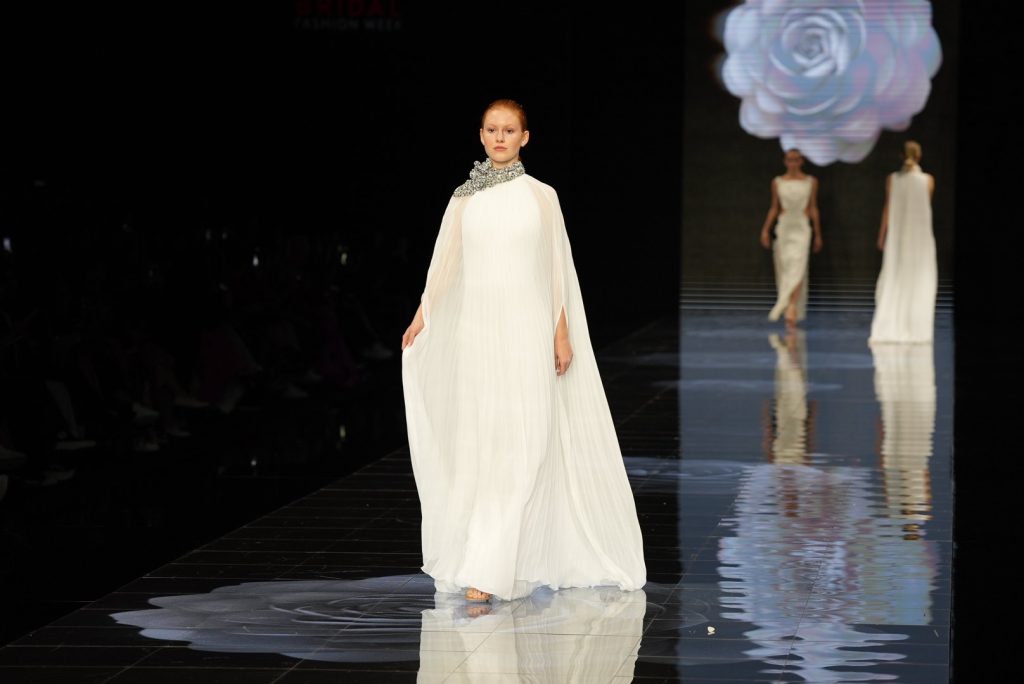In questa immagine una modella indossa un lungo abito da sposa Isabel Sanchis 2024 con un mantello impalpabile. 
