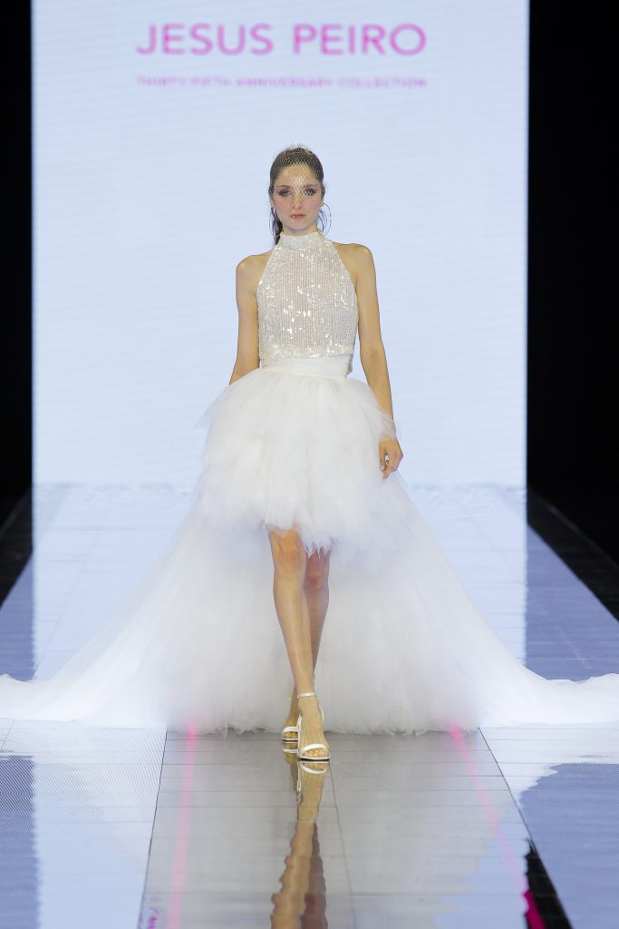 In questa immagine un abito da sposa Jesus Peiro 2024 con gonna corta in tulle che poi si allunga nella parte posteriore. 
