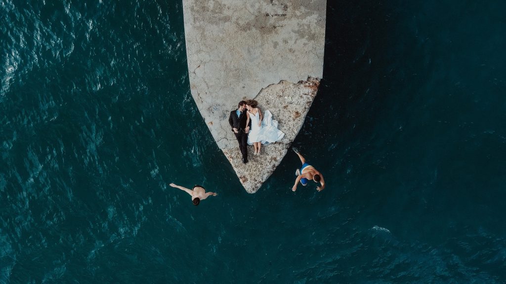 In questa foto di Mario Marinoni, tra i migliori fotografi di matrimonio a Bari, due sposi si baciano distesi su un pontile mentre altre due persone in costume si tuffano in mare