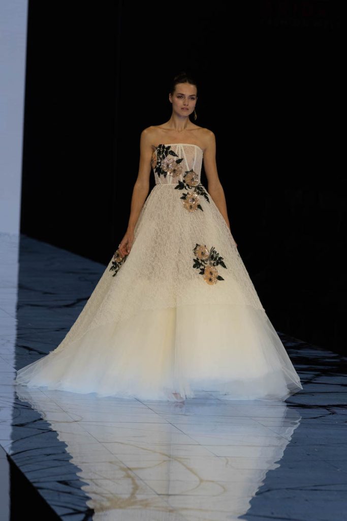 In questa foto una modella sfila indossando un abito da sposa Marco y Maria ampio con bustier e ricami di fiori colorati sul corpetto e sulle lunghezze 