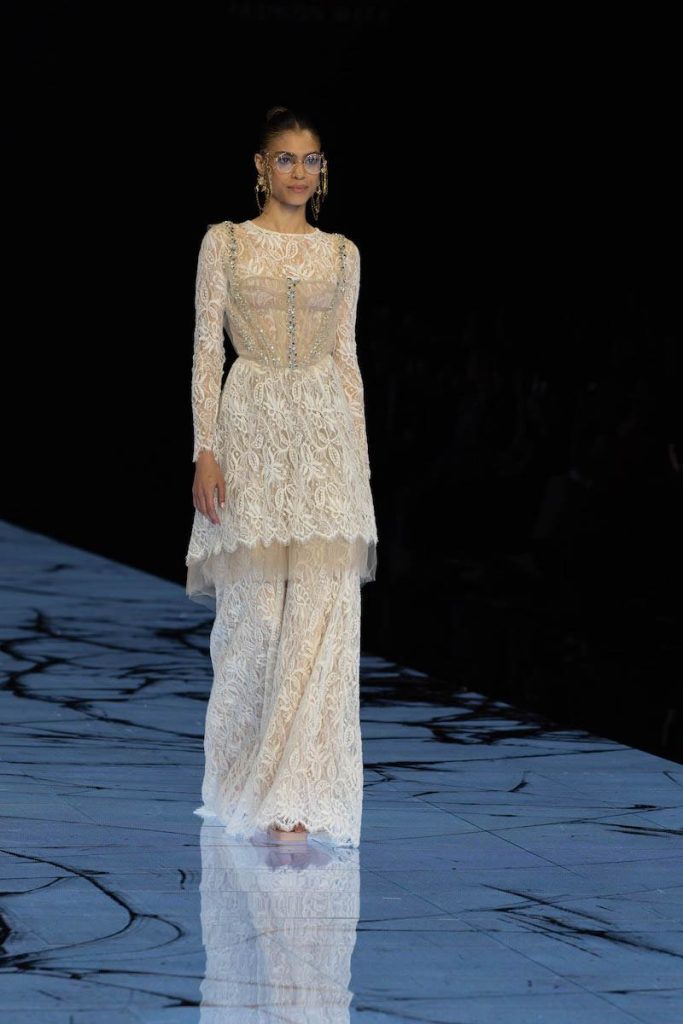 In questa foto una modella sfila indossando un abito da sposa Marco y Maria 2024 in pizzo con maniche lunghe, maxi balza e bustier in tulle