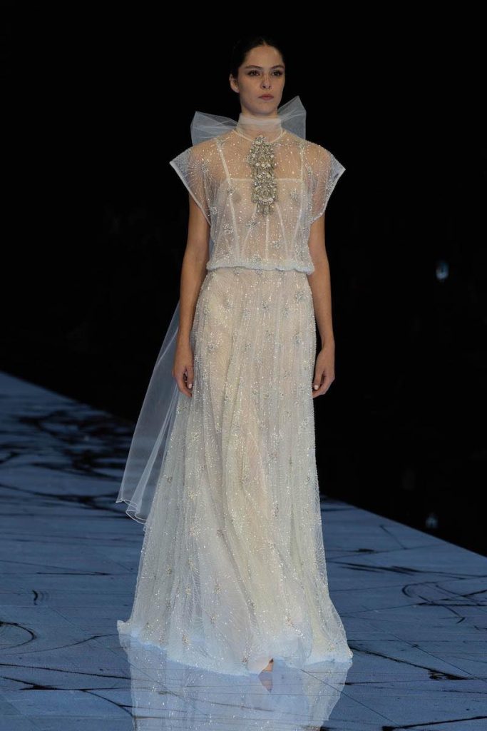 In questa foto una modella sfila indossando un abito da sposa scivolato Marco y Maria con ricami di perline e fiocco di tulle sulla nuca e dettaglio gioiello sul corpetto