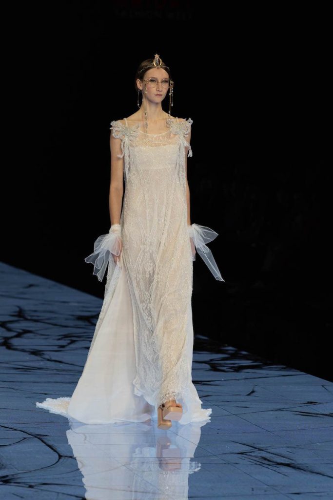 In questa foto una modella sfila indossando un abito da sposa scivolato con ricami di fiori e fiocchi di tulle sui polsi 