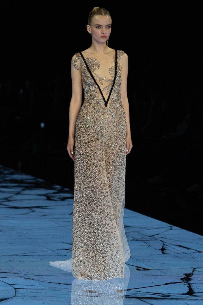 In questa foto una modella sfila indossando un abito da sposa Marco y Maria con ricami di perline all over, scollatura a V e dettaglio nero lungo lo scollo