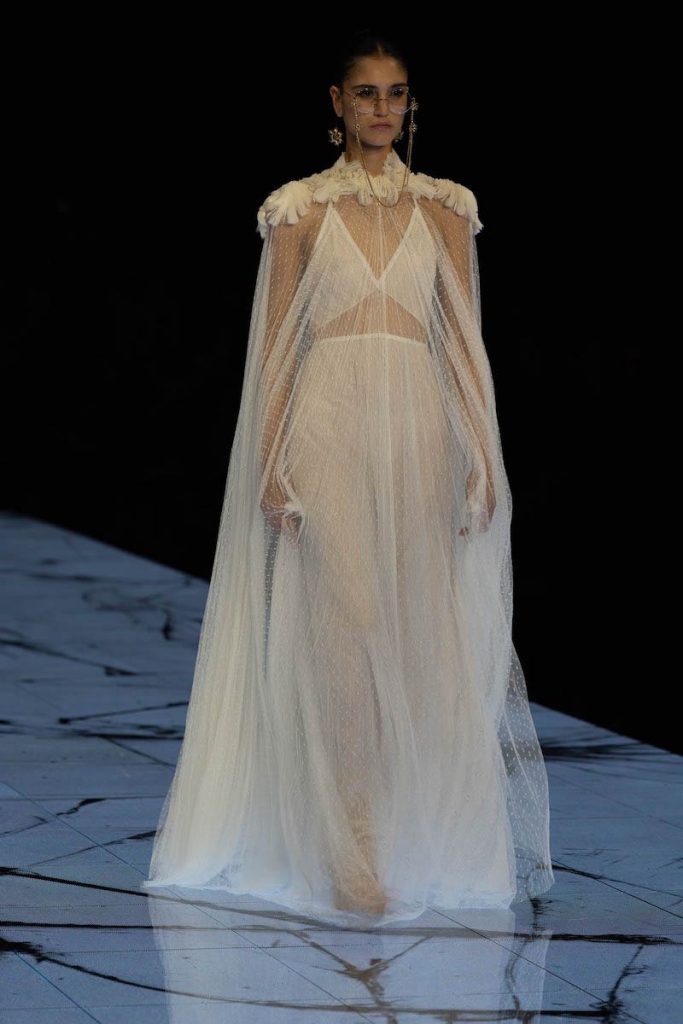 In questa foto una modella sfila indossando un abito da sposa Marco y Maria 2024 due pezzi con cappa in tulle plumetis e dettagli di rose 3D sulle spalle
