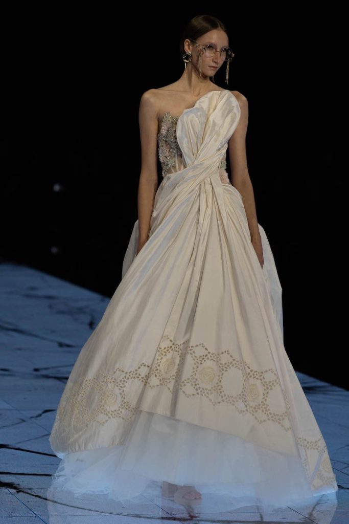 In questa foto una modella sfila indossando un abito da sposa Marco y Maria 2024 in duchesse con ricami sull'orlo e drappeggio torchon sul busto 